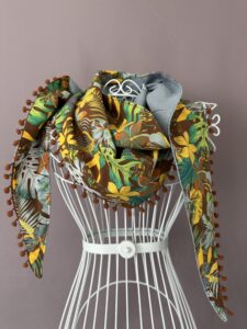 Un grand foulard triangulaire en lin et coton, réversible. Une face unie en lin gris perle, l'autre face en coton coloré imprimé avec motifs façon jungle. Le chèche est bordé de petits pompons marrons assortis au tissu.